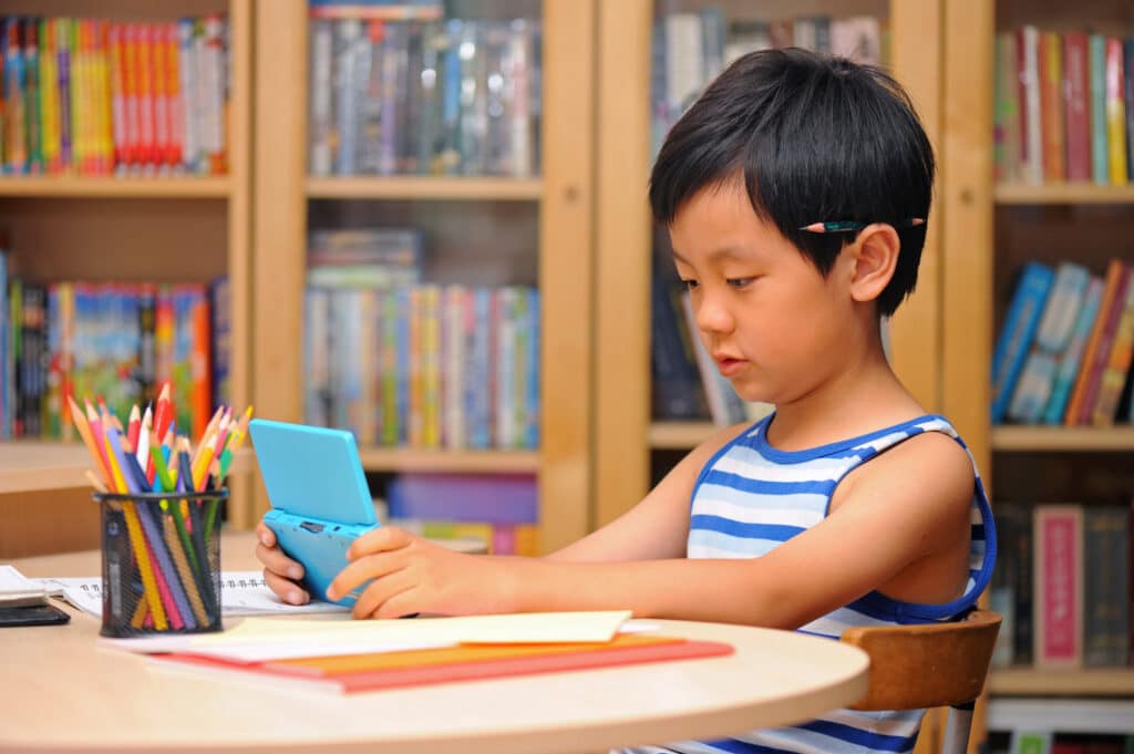 little boy playing a handheld video game instead of coloring with pencils on table