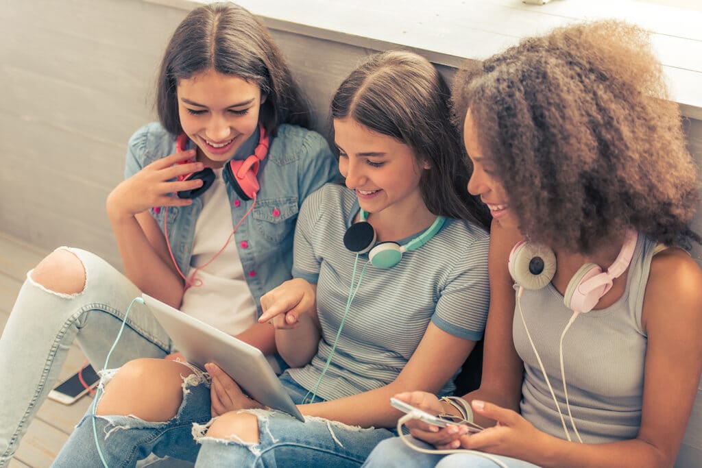 three teen girls smiling with headphones around their necks looking at an ipad